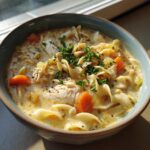 Close-up of a bowl of creamy chicken noodle soup with carrots, noodles, and herbs.