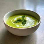 A bowl of creamy pea and mint soup, garnished with mint leaves and a swirl of cream.