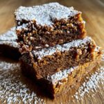 A close-up of three stacked Gingerbread Cookie Bars dusted with powdered sugar on a wooden surface.