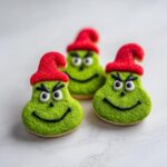 Three festive Grinch cookies decorated with green sugar, red hats, and black icing smiles and eyes.