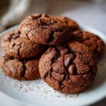 A close-up of a stack of rich, dark Hot Cocoa Cookies generously studded with chocolate chips and dusted with cocoa powder.