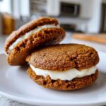 Two Incredible Gingerbread Oatmeal Cream Pies stacked on a white plate, filled with creamy white frosting.