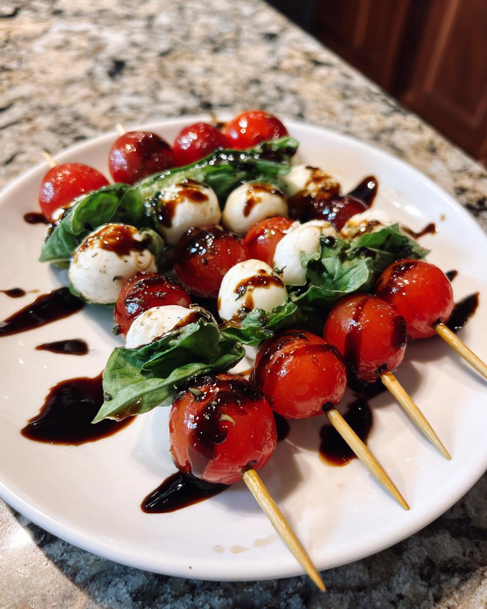 A white plate filled with Mini Caprese Skewers featuring cherry tomatoes, mozzarella balls, basil, and drizzled with balsamic glaze.