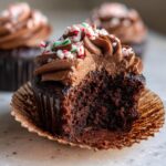 A close-up of a decadent Peppermint Mocha Cupcake with rich chocolate frosting and crushed candy canes, with a bite taken out.