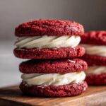 Close-up of stacked Red Velvet Cookies with creamy white frosting on a wooden board.
