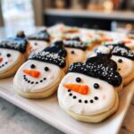 Close-up of adorable snowman hat cookies with white icing, black hats, orange noses, and black dot smiles.