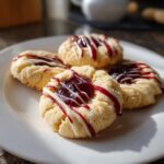 Close-up of white chocolate raspberry thumbprint cookies on a white plate. The cookies are drizzled with white chocolate and raspberry jam.